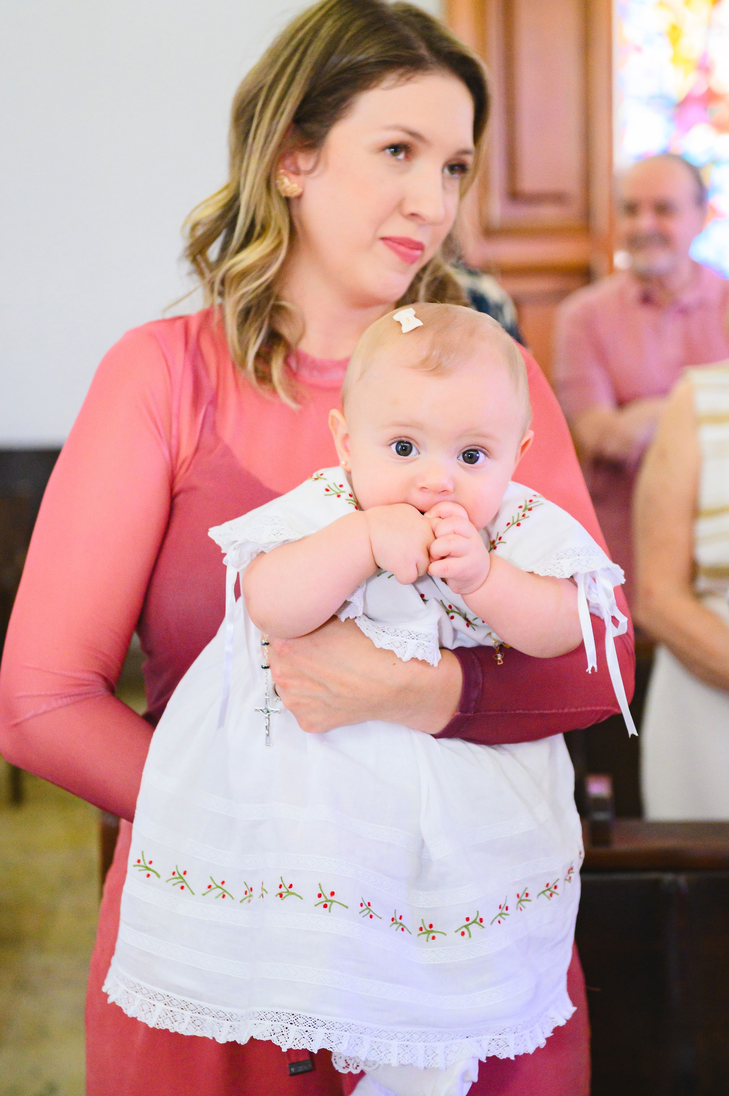 A baby in a white christening gown is held while chewing on their hand during a special ceremony.