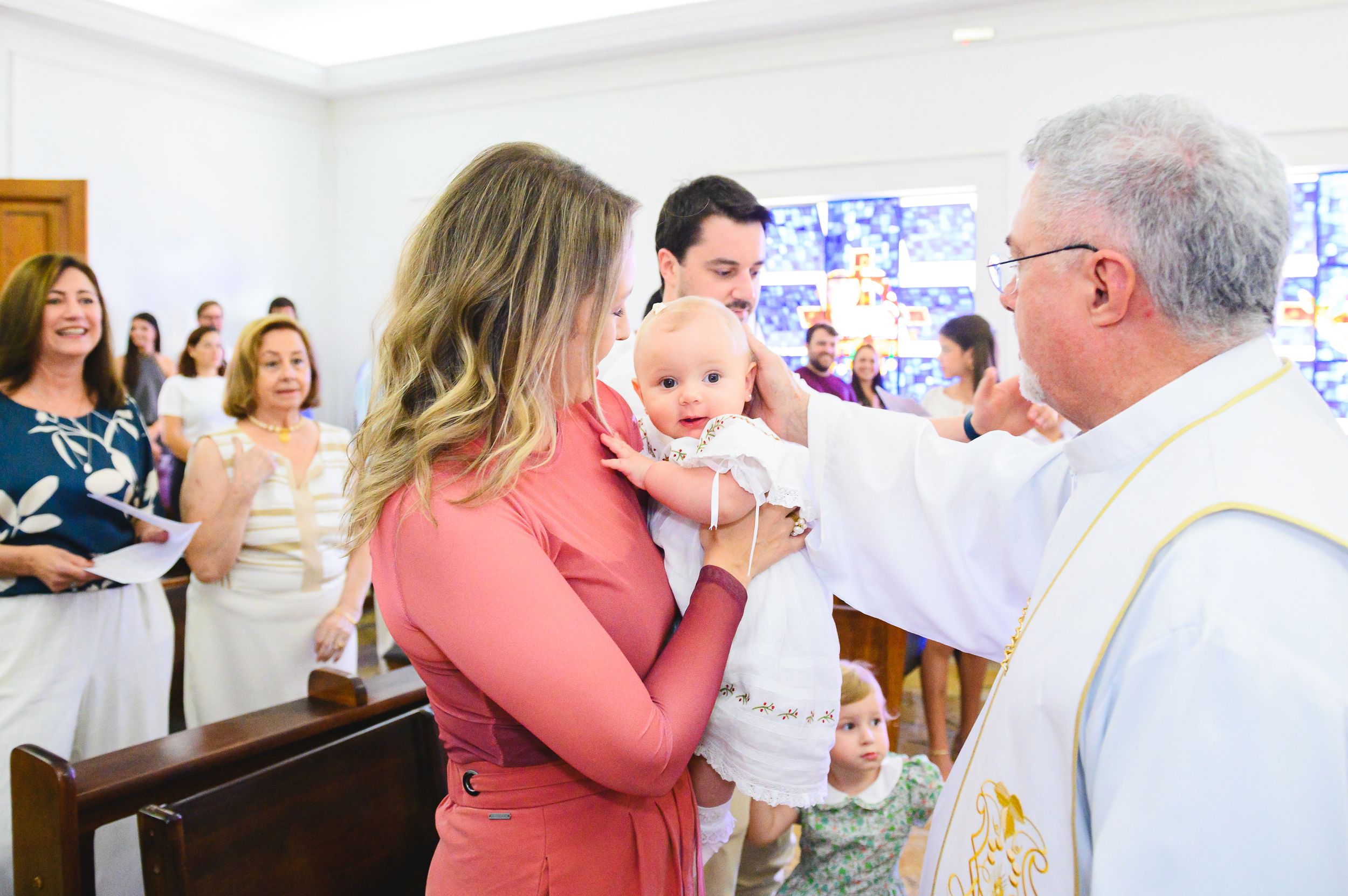 A religious ceremony takes place in a church as a priest in white robes performs a blessing ritual with gathered guests.