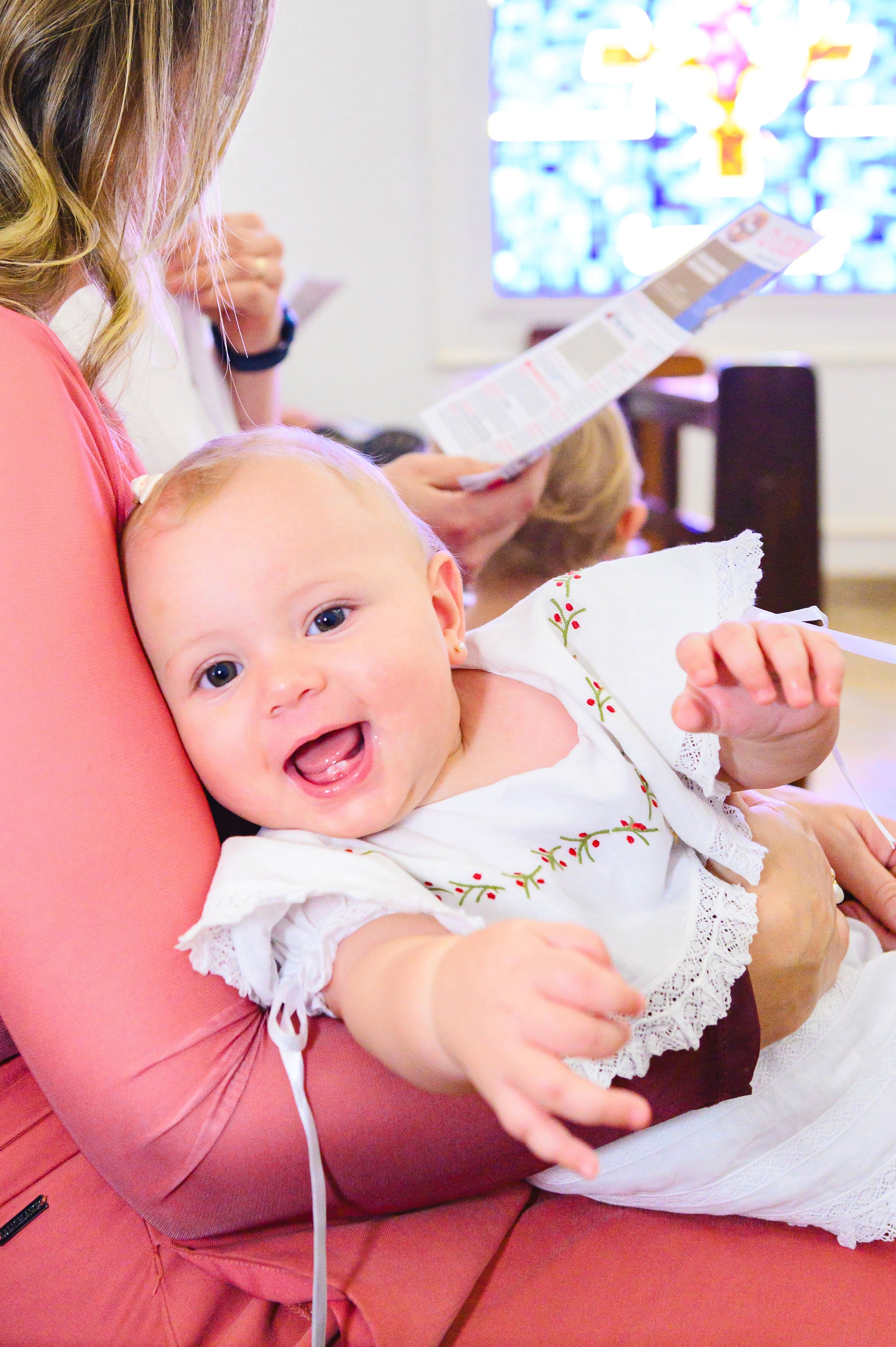 A happy baby wearing a white embroidered dress sits in someone's lap while being read to.
