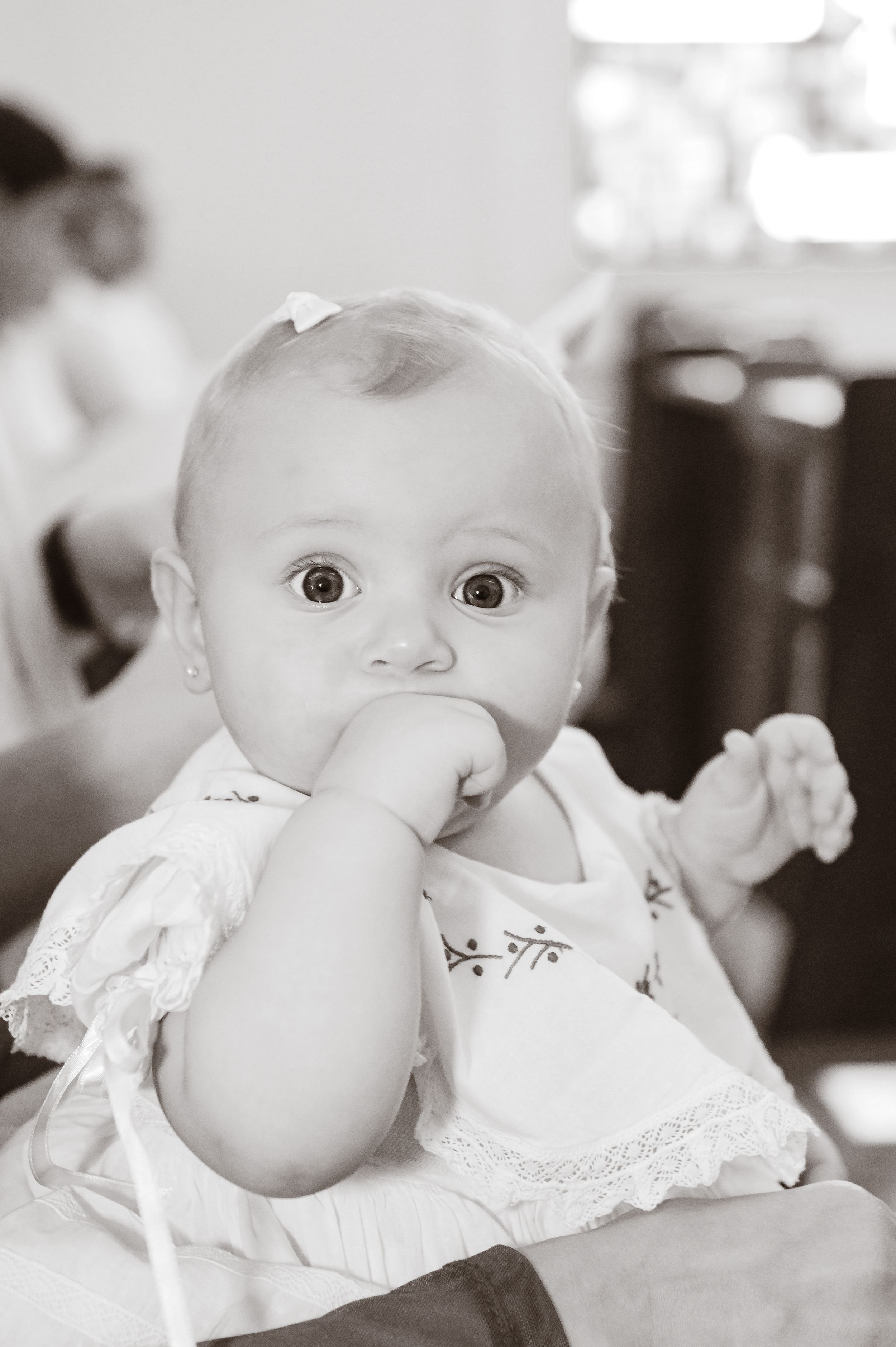 Black and white portrait of a baby in white clothing with wide eyes and fingers in their mouth.