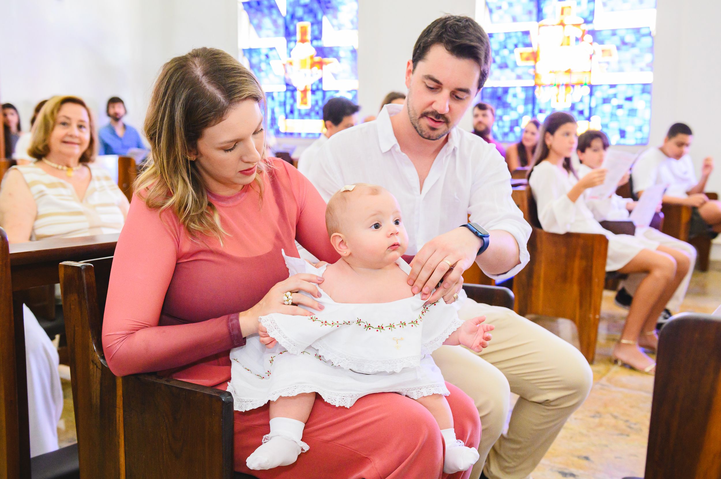A baby in white clothing sits between two people in a church pew during a baptism ceremony.