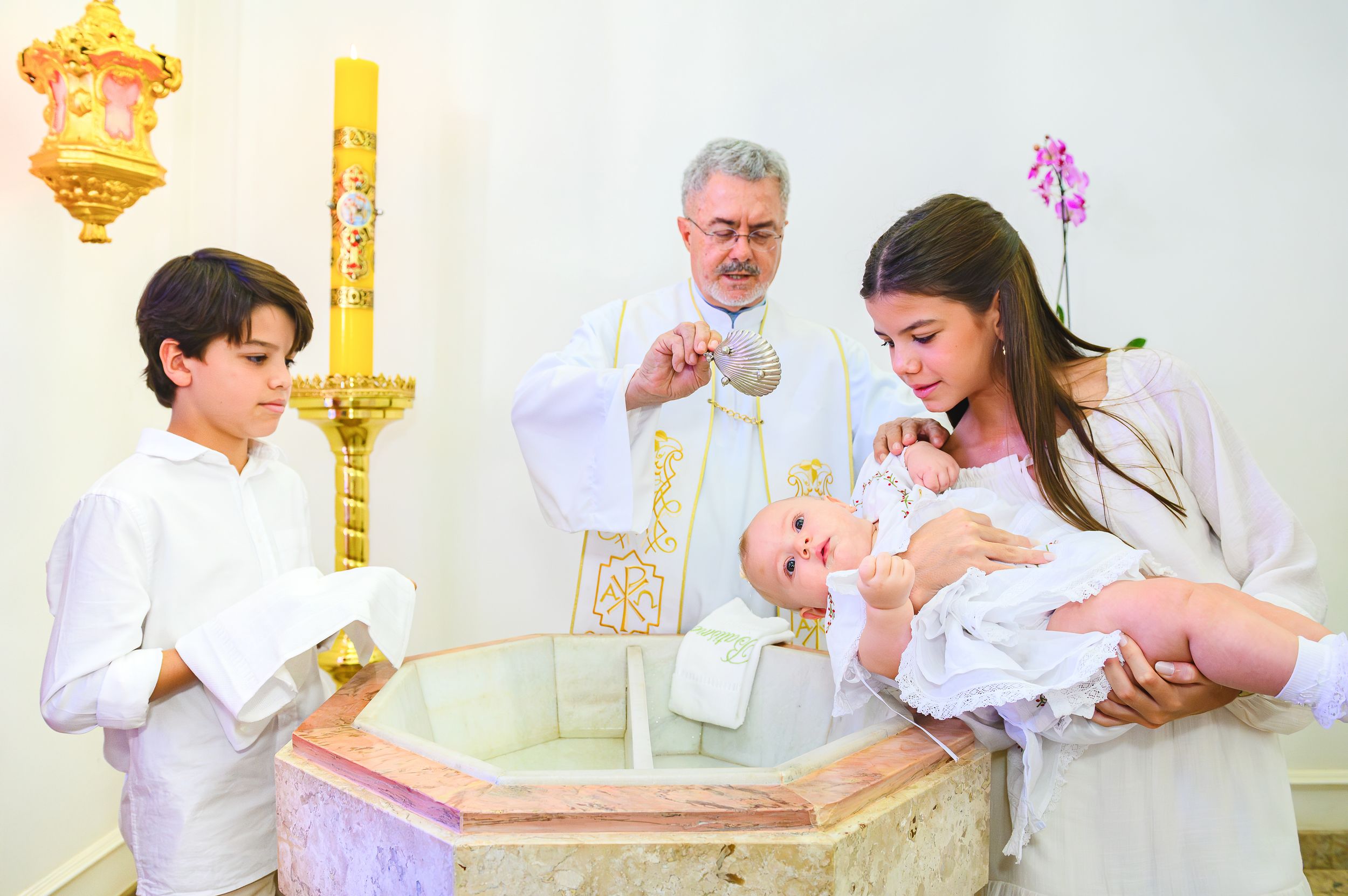 A religious baptism ceremony takes place at a marble baptismal font with clergy and family members present.