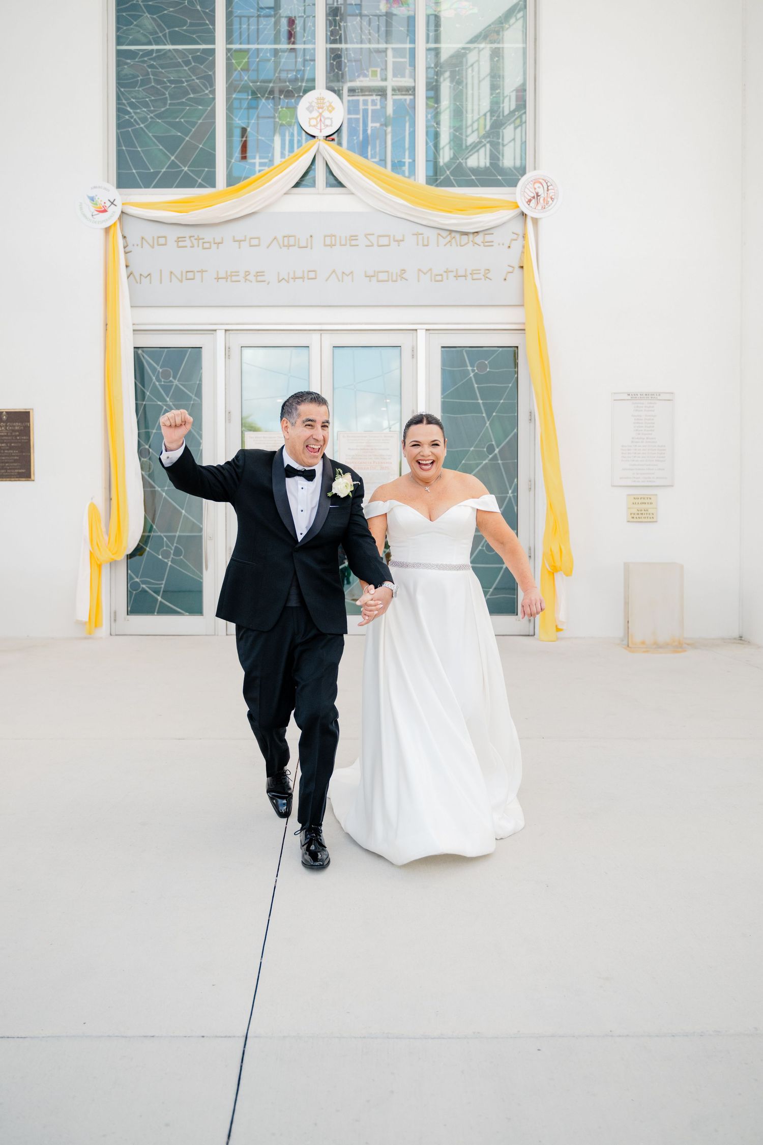 A newlywed couple celebrates with joy as they exit a church ceremony under a yellow and white draped archway.