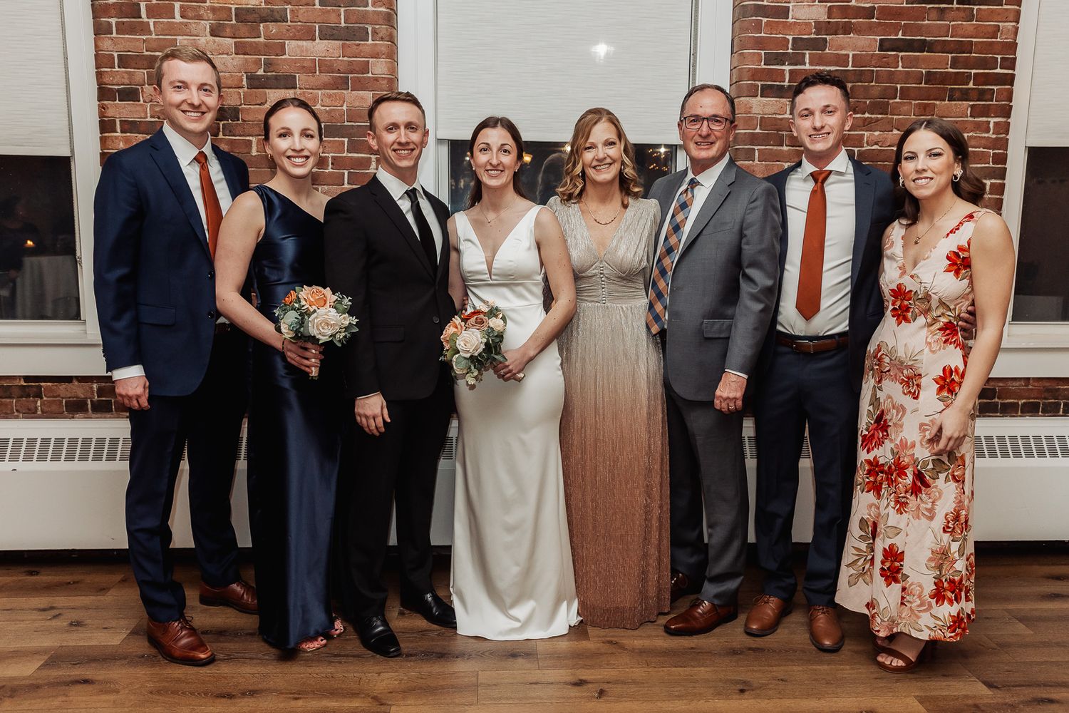 Wedding party in formal attire poses together against brick wall at indoor reception venue.