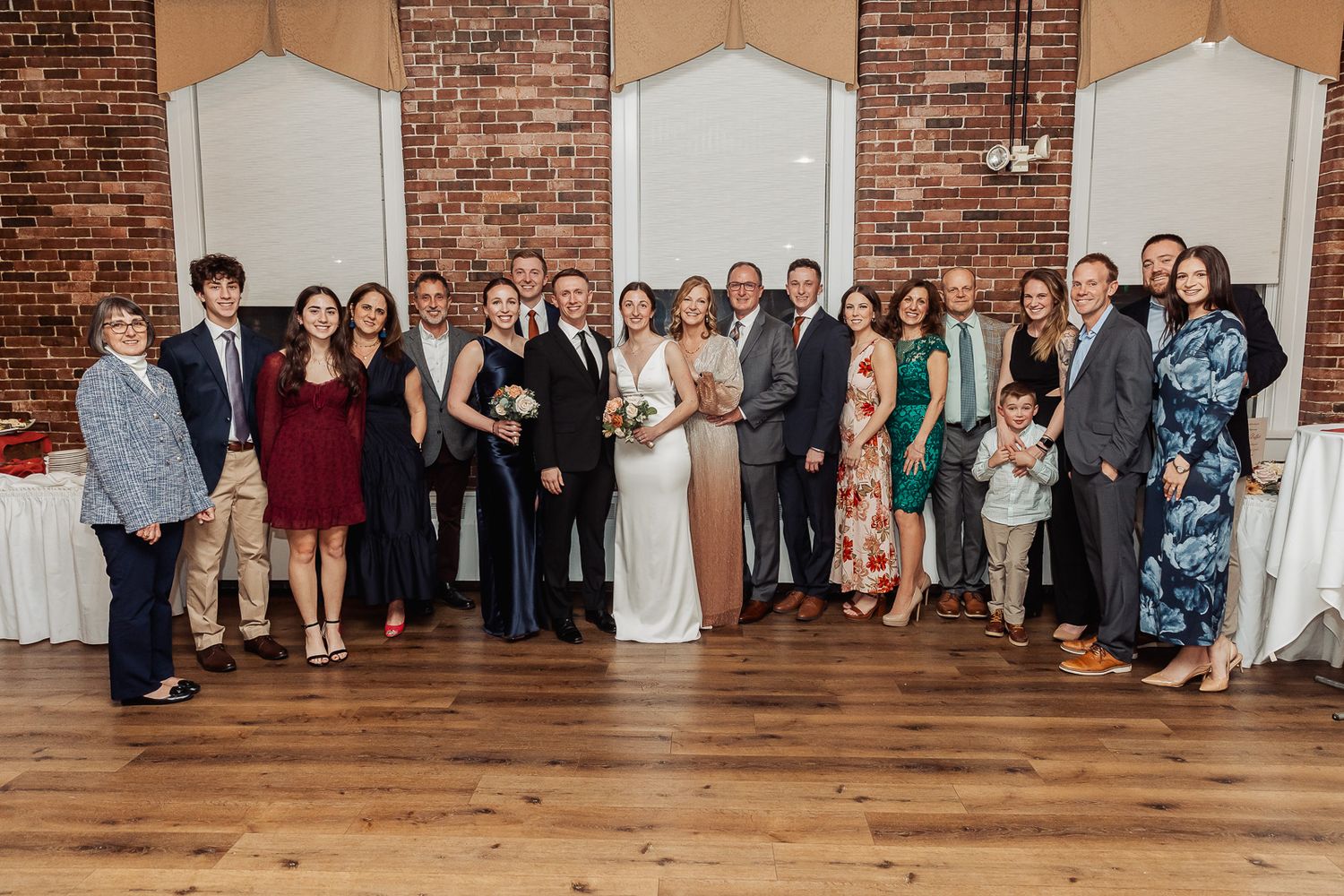 Large wedding party poses together in formal attire against exposed brick wall at indoor reception venue.
