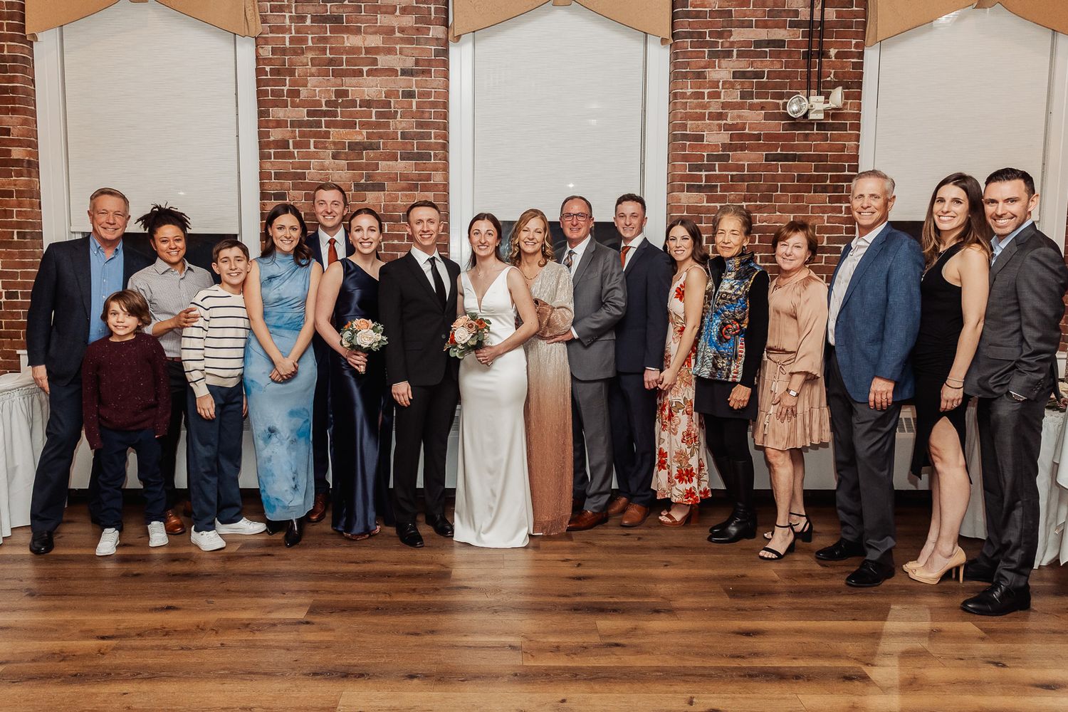 Large formal group photo of wedding party and guests standing together on wooden floor in front of brick wall.