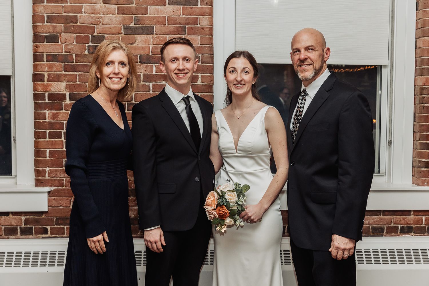 Four people in formal attire pose for a photo against a brick wall at a wedding ceremony.