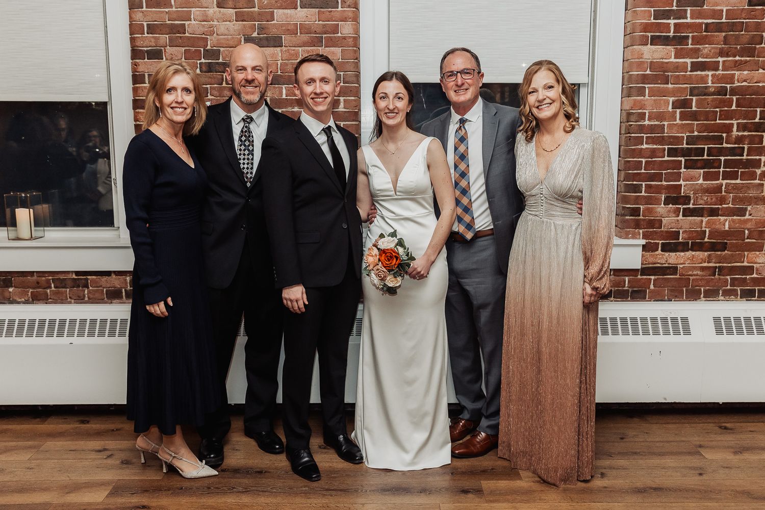 A group of six people stand together at a wedding celebration against a brick wall backdrop.