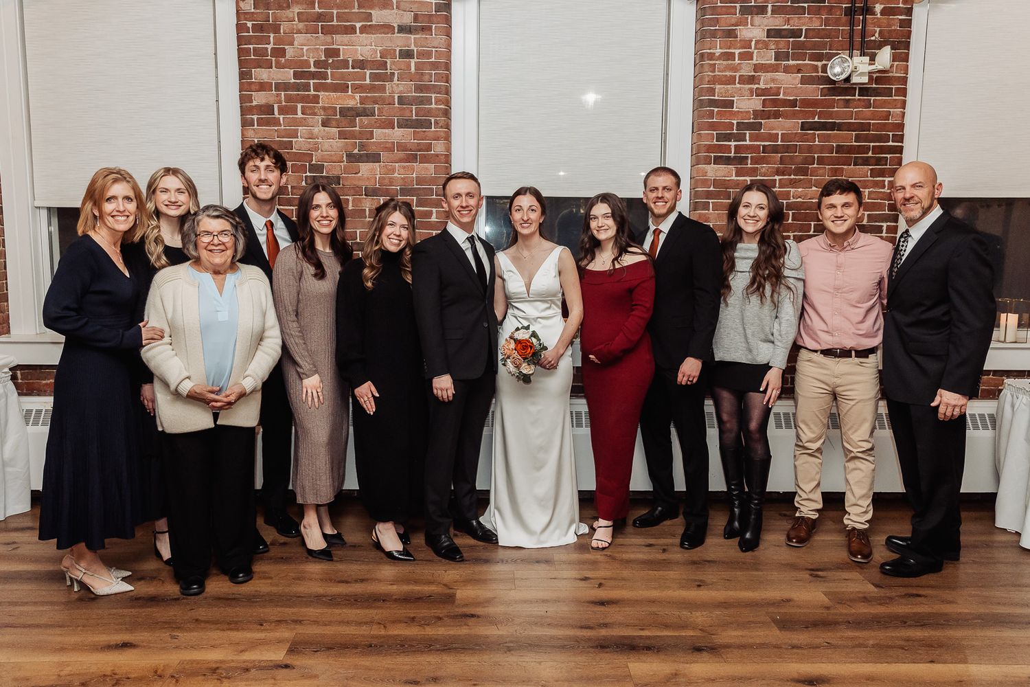 A wedding party poses together in formal attire against a brick wall backdrop in an indoor venue.
