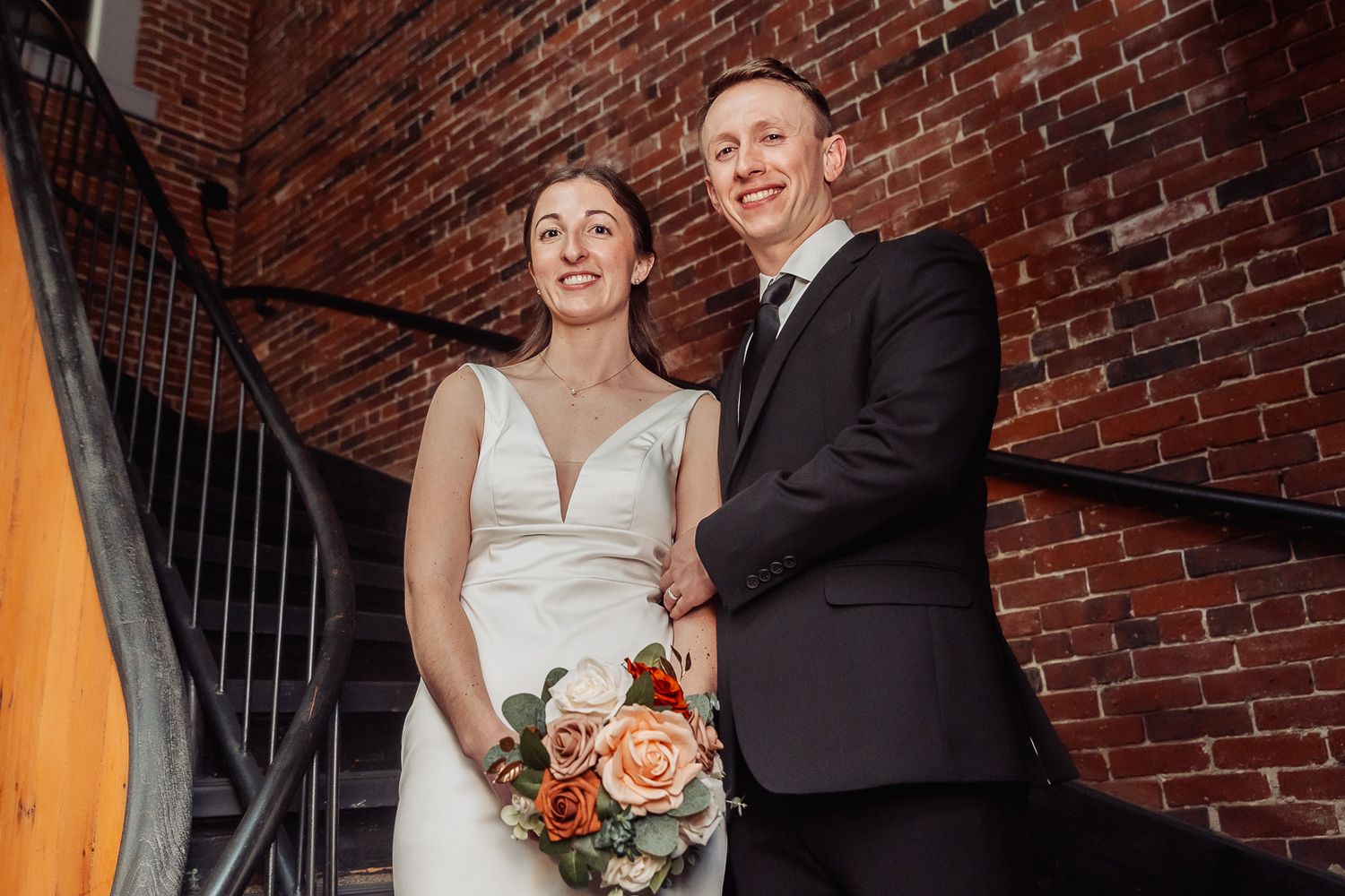 Wedding couple smiles while standing on metal staircase against red brick wall with bride holding coral bouquet.