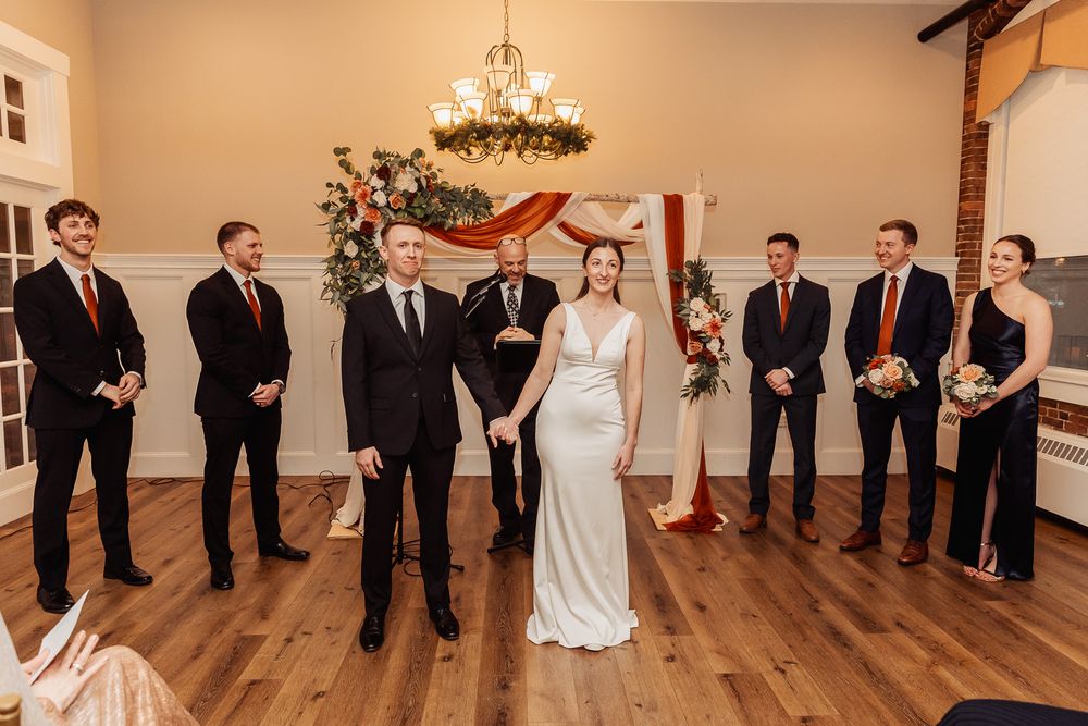 Wedding party group poses under decorative chandelier on hardwood floor in rustic indoor venue.
