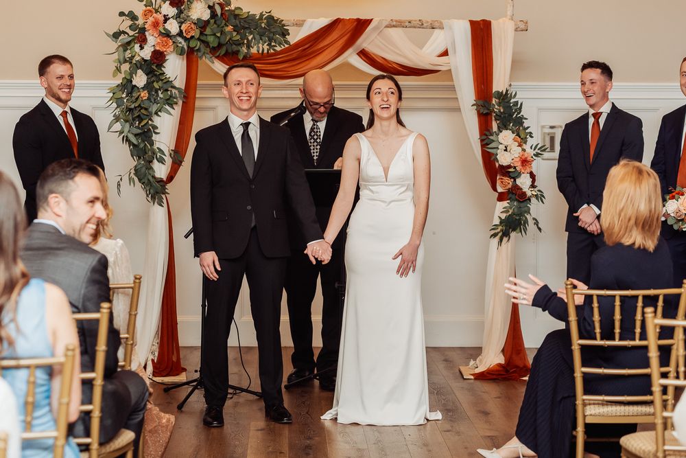 Wedding ceremony takes place in elegant indoor venue with copper arch and floral decorations.