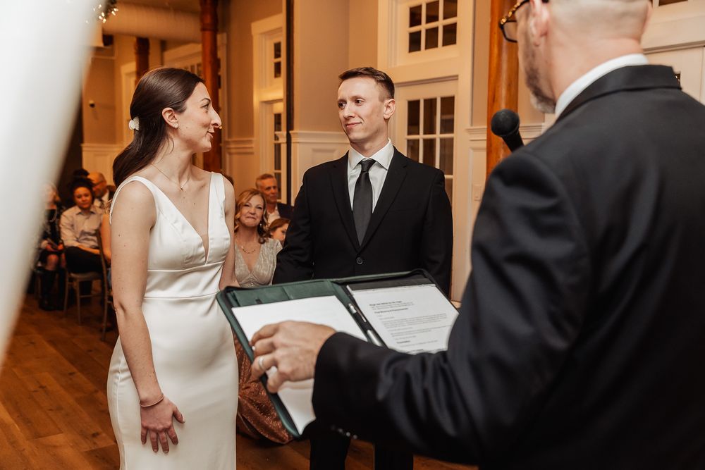 A couple stands before an officiant during an indoor wedding ceremony in an elegant venue with wooden floors.