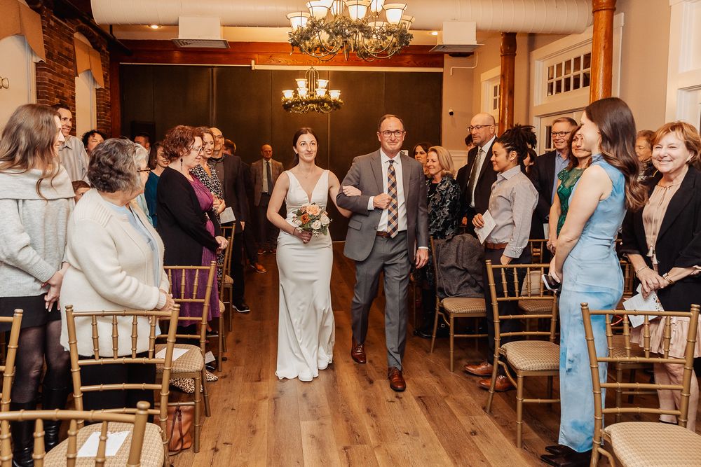 A couple walks down a wooden aisle during their wedding ceremony while guests watch from elegant wooden chairs under chandeliers.