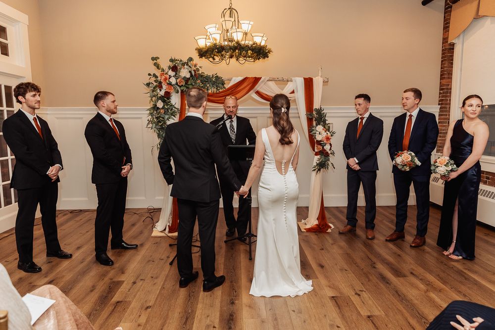 Wedding ceremony inside a rustic venue with wooden floors and chandelier as wedding party gathers around couple.