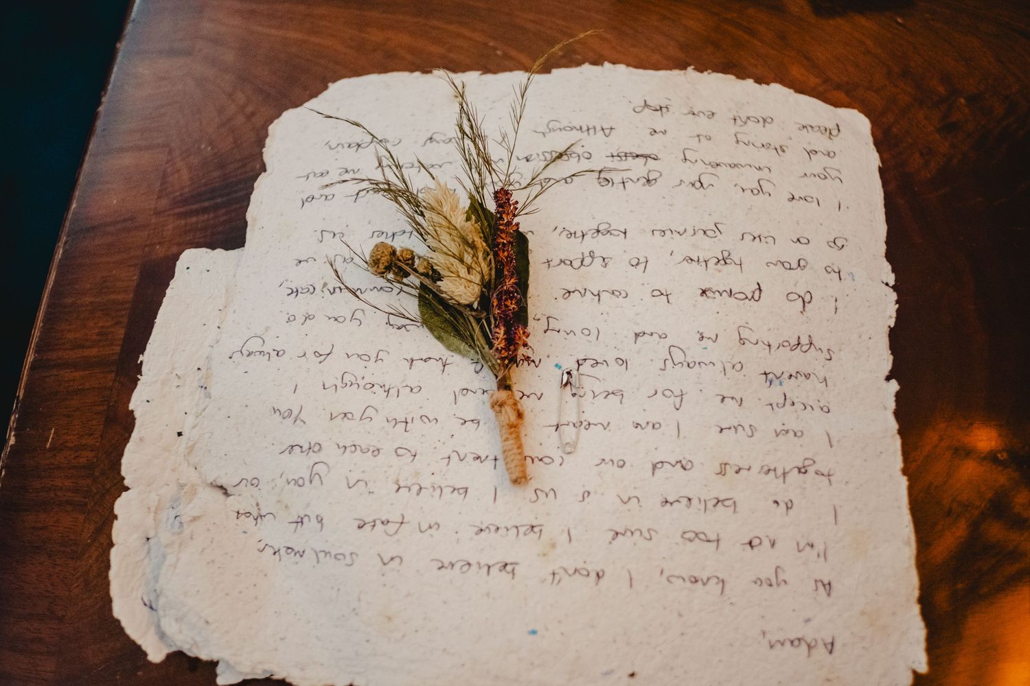 A dried flower stem rests on handwritten text on weathered, deckle-edged paper against a dark wooden surface.