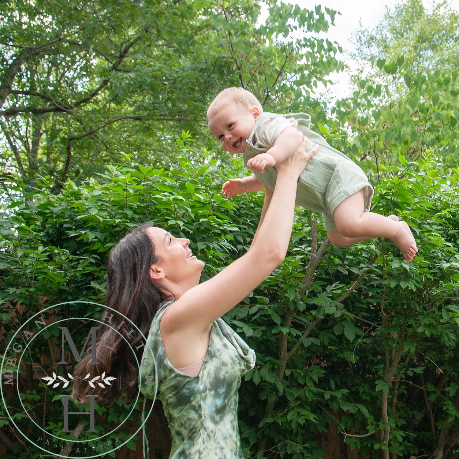 A joyful outdoor photo of mom swinging her toddler up into the air captured in a lush green garden setting on a bright summer day.