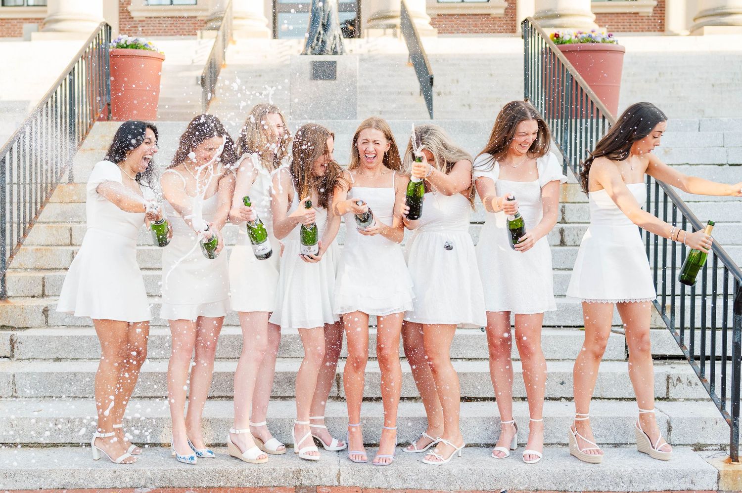 Bridesmaids in short white dresses holding bouquets stand together on stone steps while laughing and smiling.