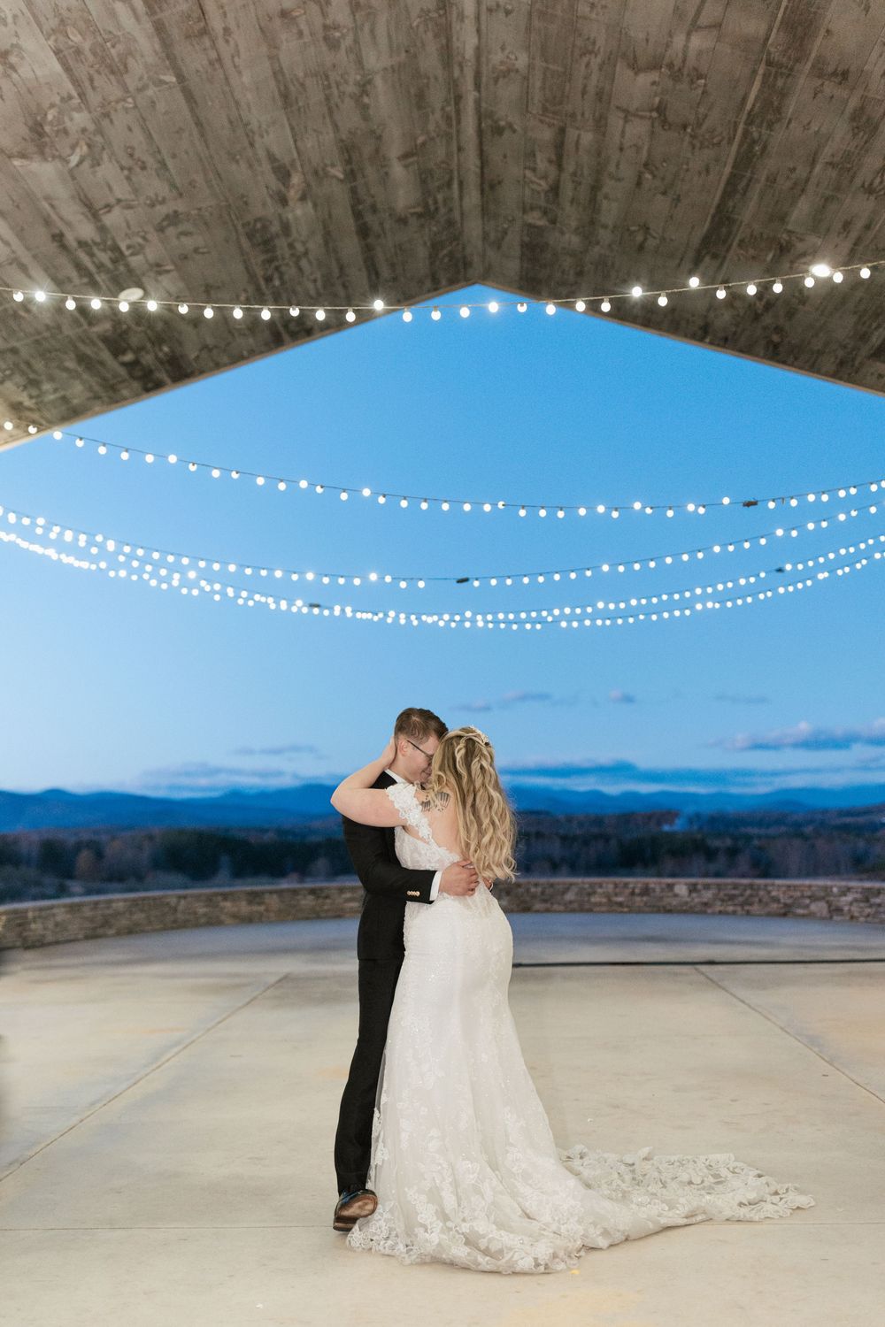 Illuminated string lights hang from a concrete ceiling over a covered outdoor terrace with mountain views.