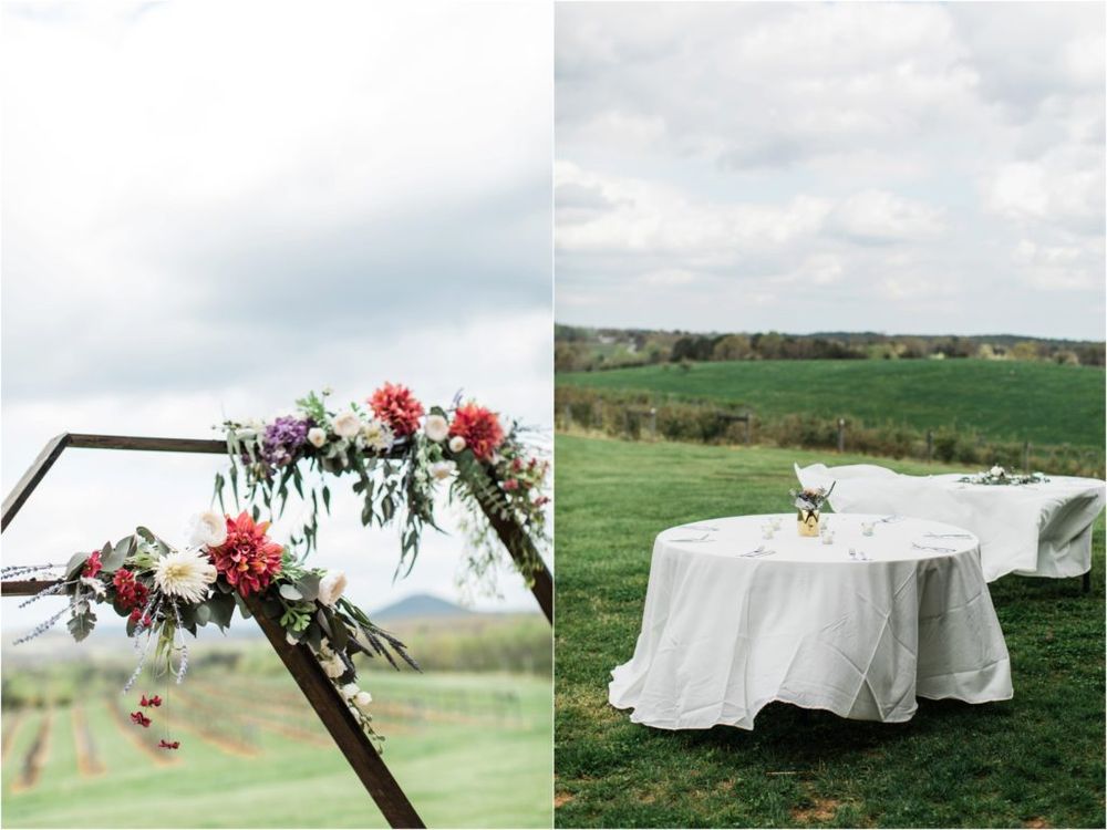 Outdoor wedding venue setup featuring a wooden arch adorned with red flowers and a white tablecloth covered table in a meadow.