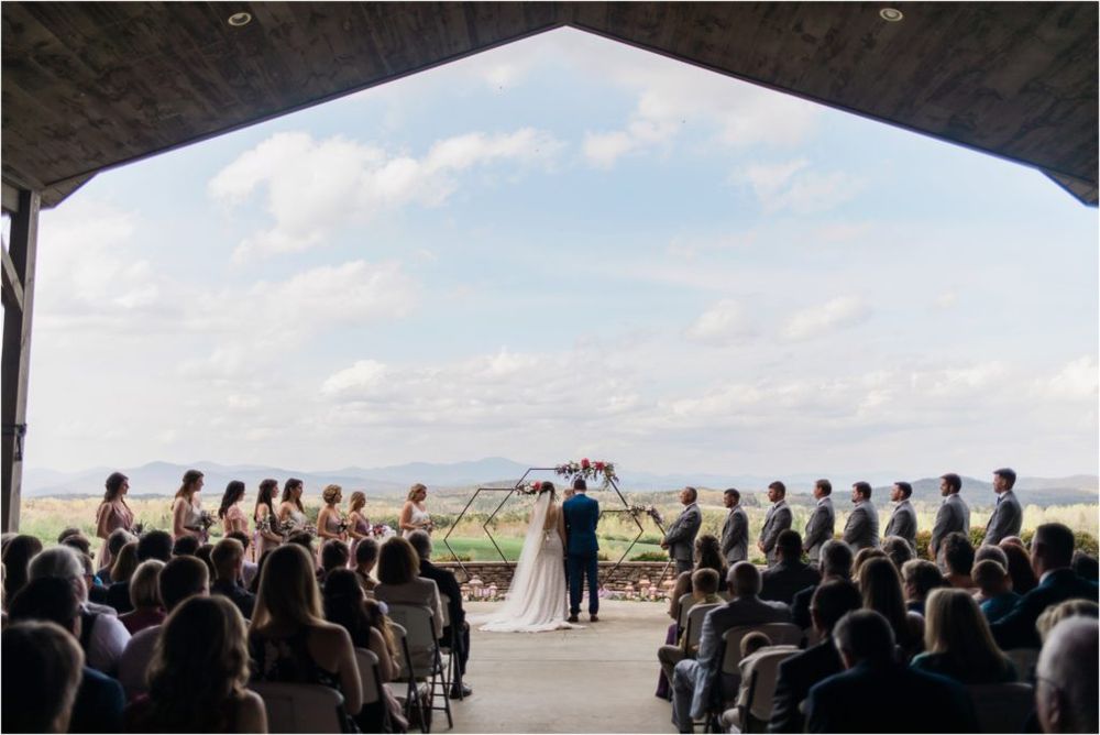 A wedding ceremony takes place under a rustic wooden arch overlooking a waterfront view with guests seated on both sides.