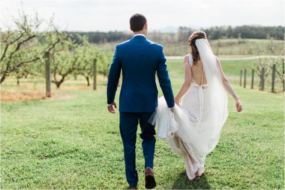 A couple on their wedding day walk through a scenic orchard with rolling hills in the background.