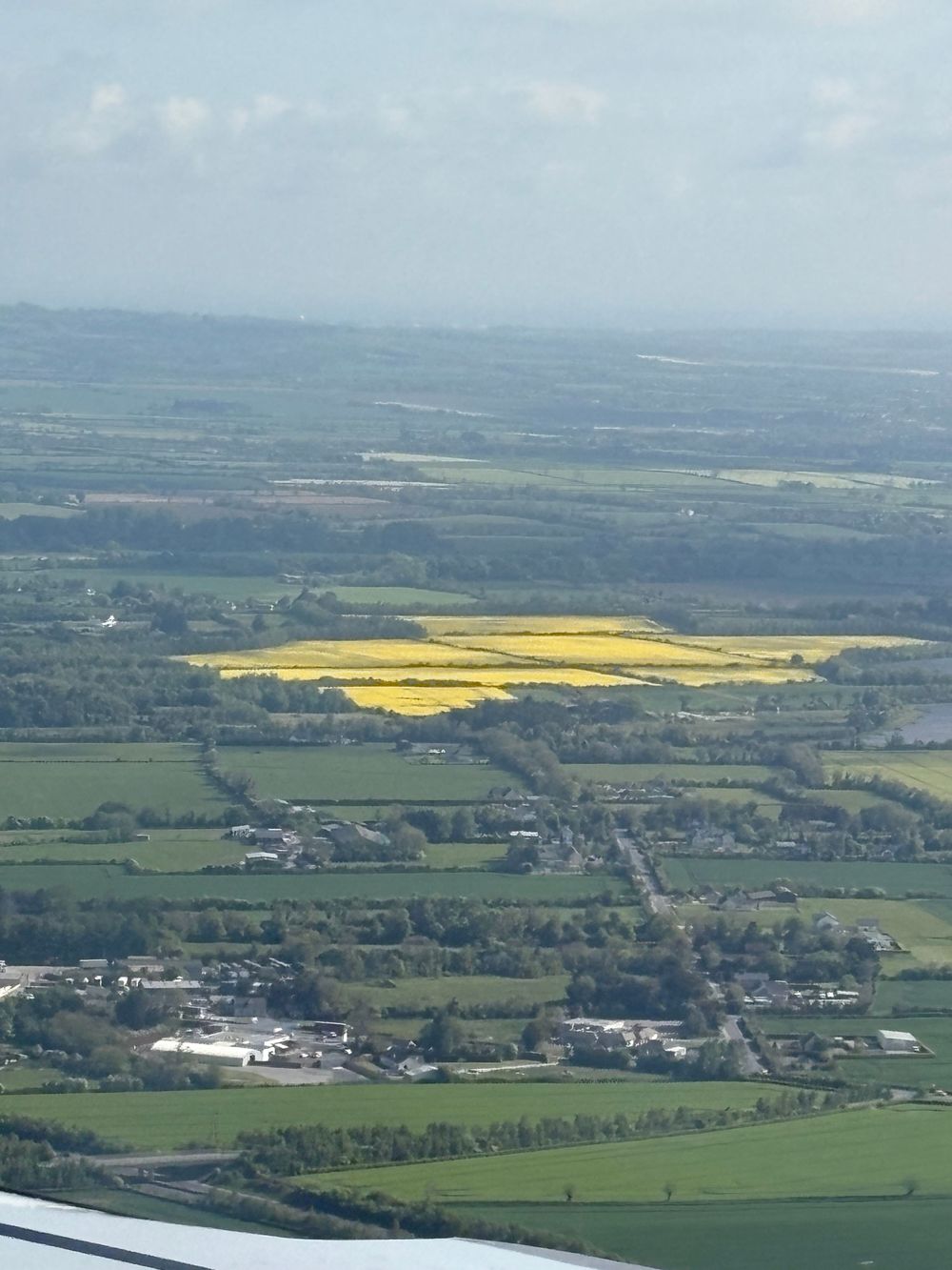 Aerial view of rural countryside with bright yellow canola fields and scattered buildings amid green farmland.