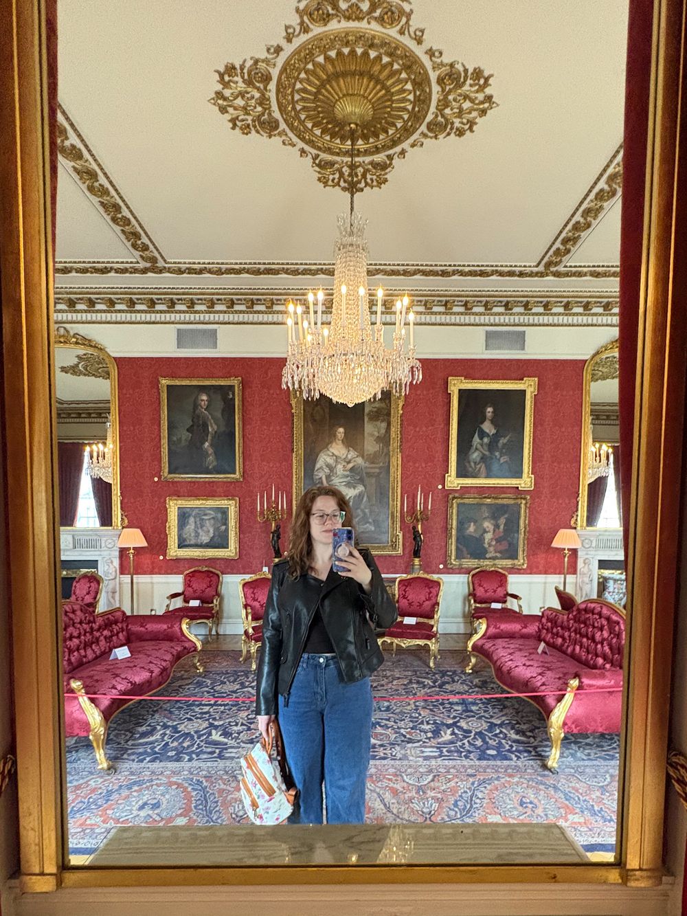Mirror selfie in an ornate historic room with red walls, crystal chandelier, and period portraits in gilded frames.