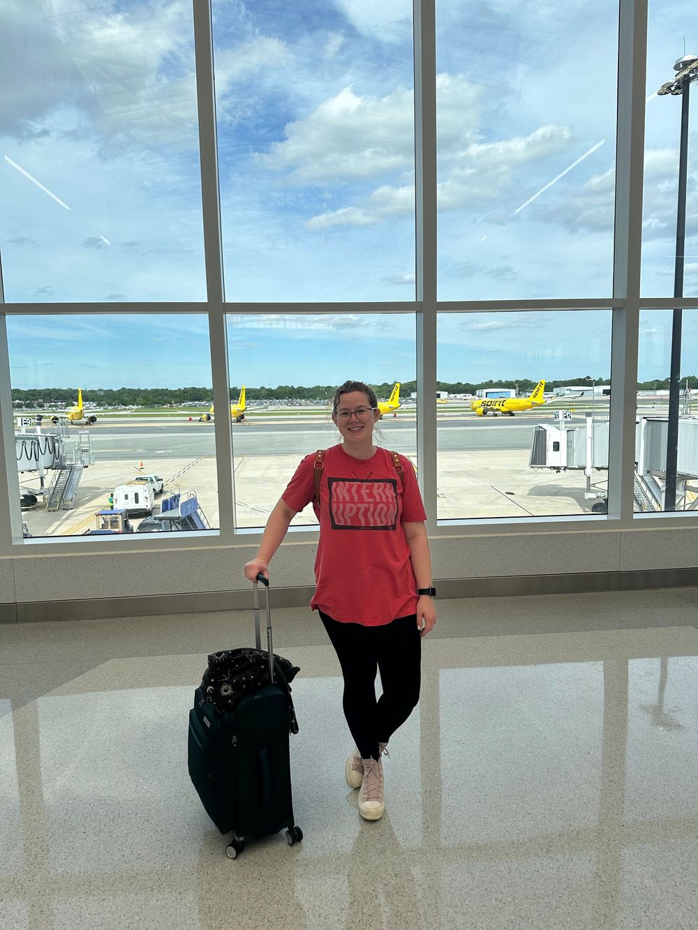 Traveler stands at airport terminal window overlooking tarmac with yellow planes and clear blue sky visible outside.
