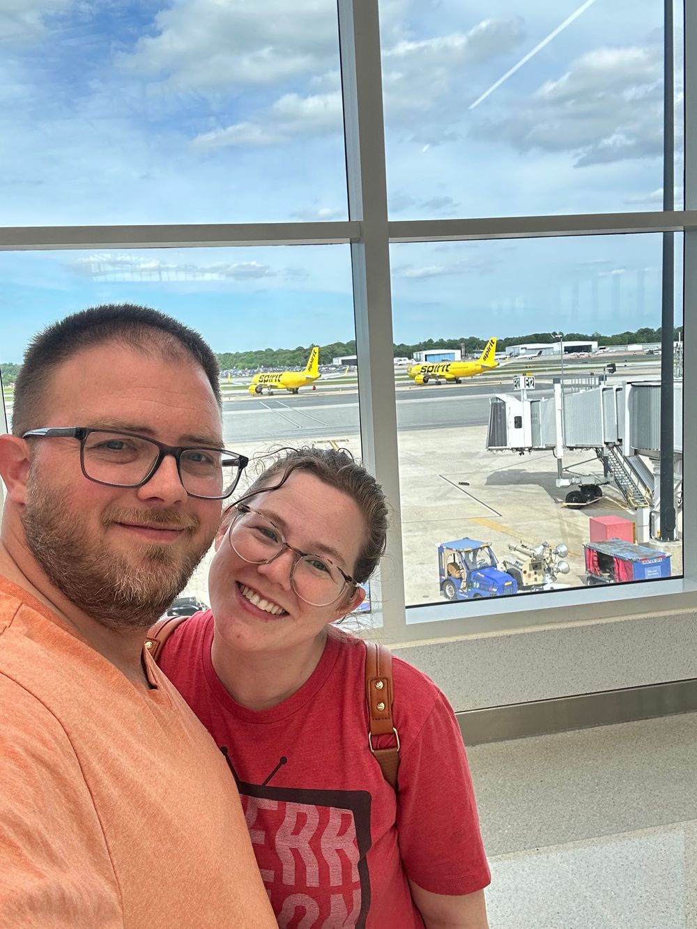 Selfie taken at an airport terminal window with yellow aircraft and jetbridge visible on the tarmac in the background.