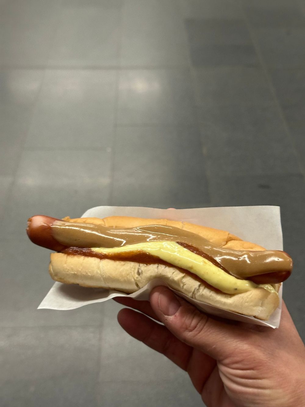 A hand holds a classic hot dog with yellow mustard and toppings on a white paper wrapper.
