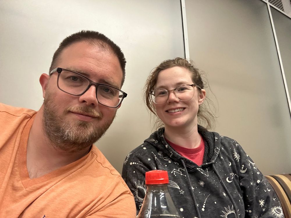 A selfie of two people wearing glasses, one in an orange shirt and one in a patterned black shirt, in an indoor setting.