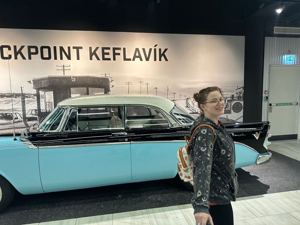 A vintage two-tone blue and white classic car is displayed at Checkpoint Keflavik with a visitor standing nearby.