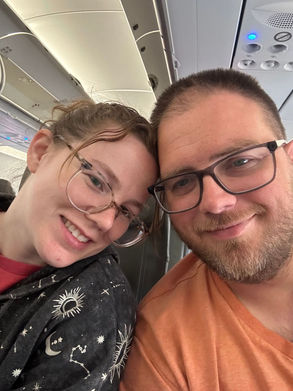 Two people wearing glasses seated next to each other on an airplane taking a selfie.