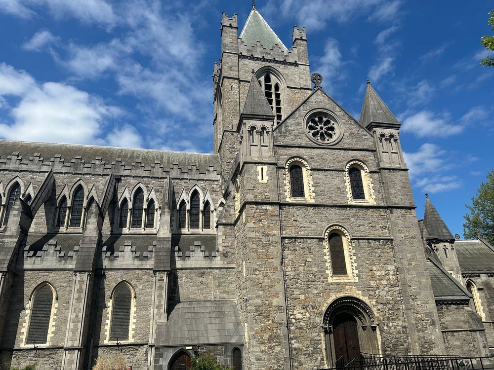 Gothic stone church exterior with pointed arches, clock tower and ornate architectural details against blue sky.