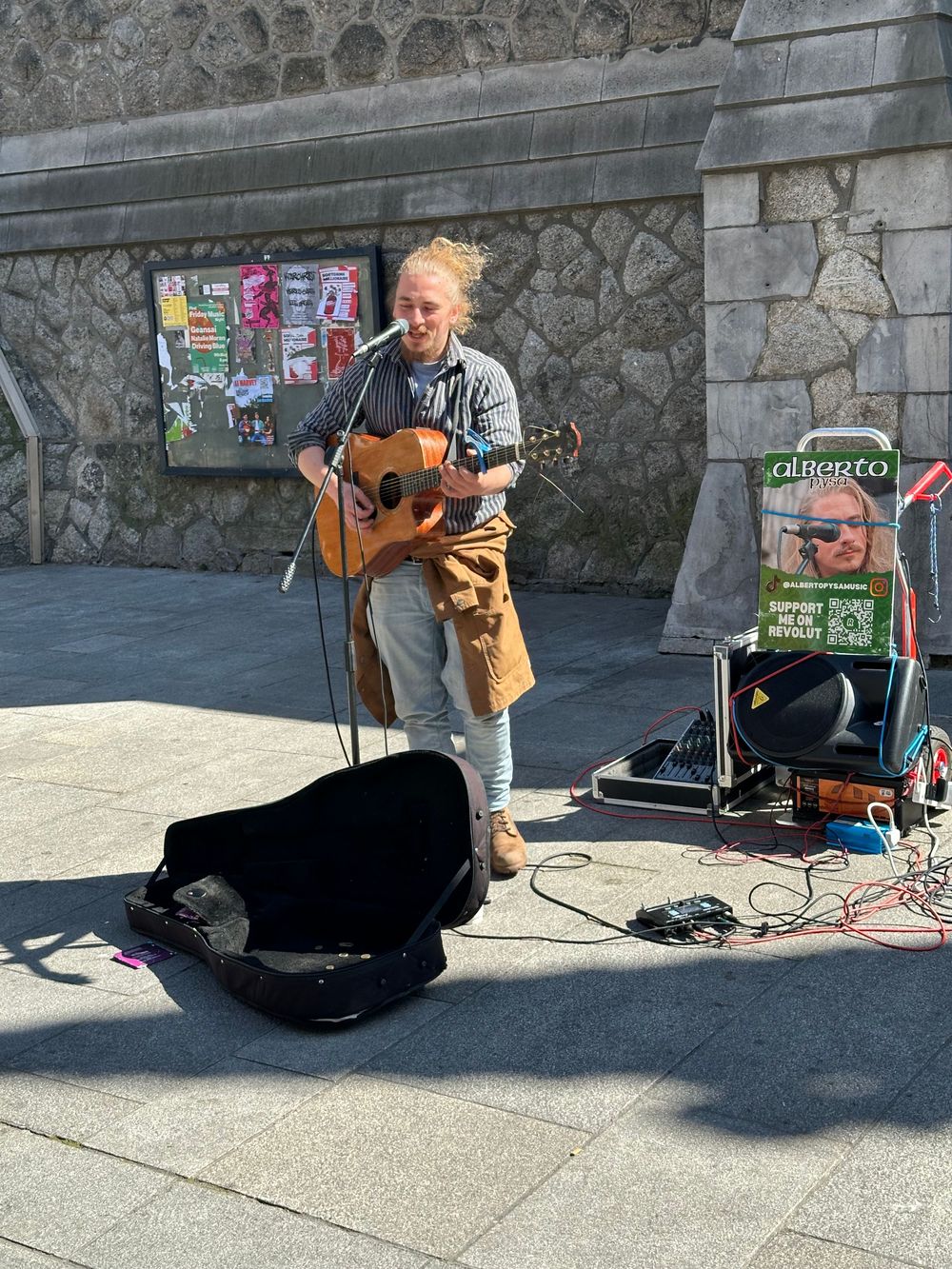 A street musician performs with an acoustic guitar and amplifier setup on a sidewalk next to an open instrument case.