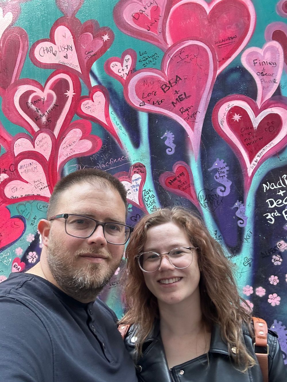 A couple poses together in front of a colorful heart-themed mural with pink and red hearts on a teal background.