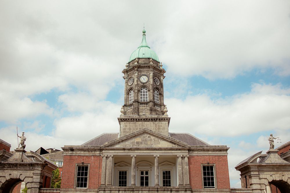 Historic Dublin Castle with neoclassical architecture features a prominent clock tower and copper dome against cloudy Irish skies.