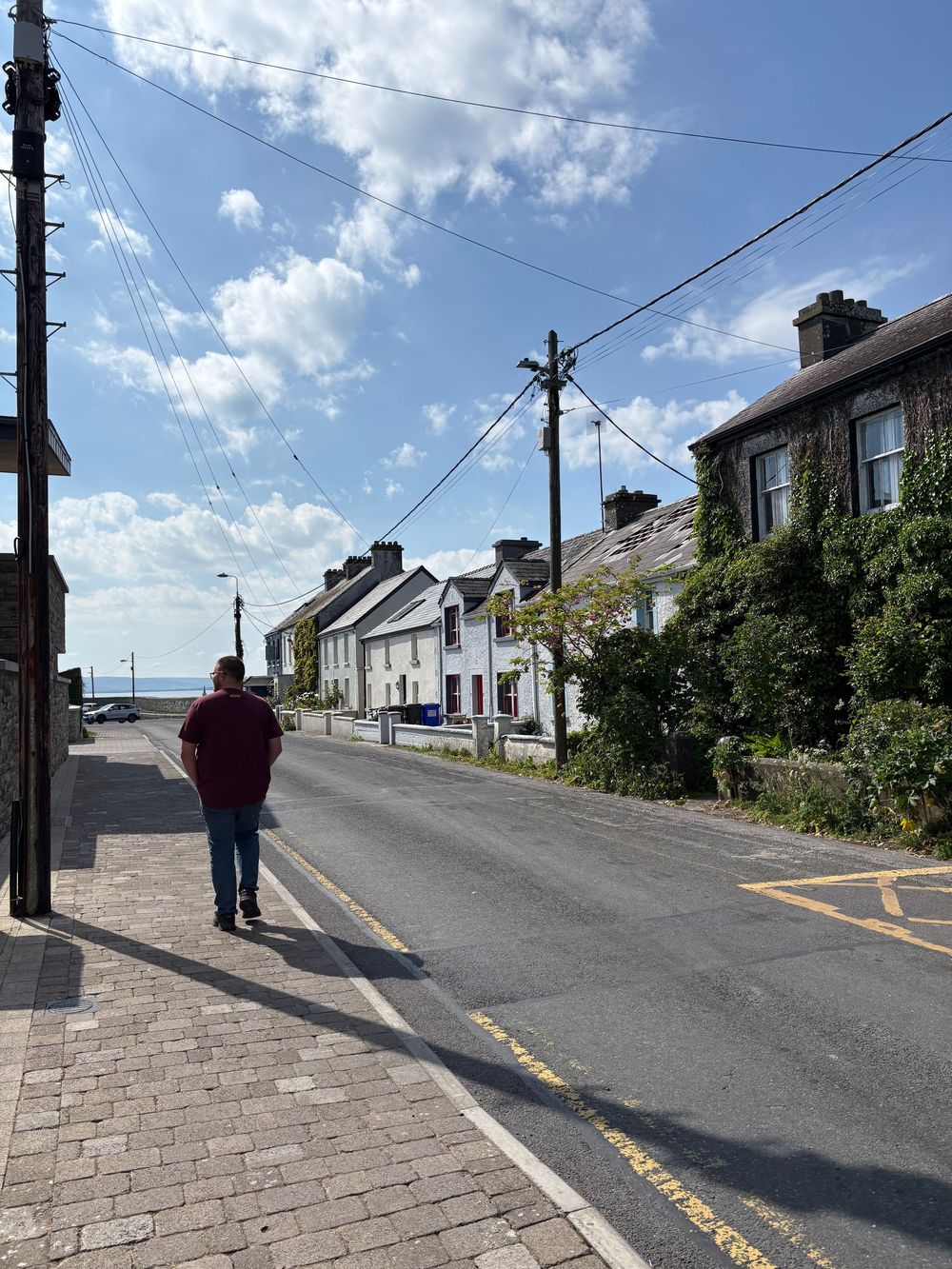 A person walks along a quiet coastal street lined with white houses and power lines under a blue cloudy sky.