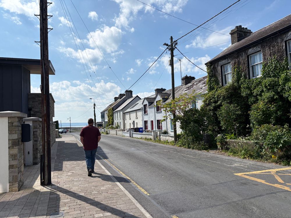 Person walking along a quiet village street with whitewashed houses and utility poles under a blue summer sky.