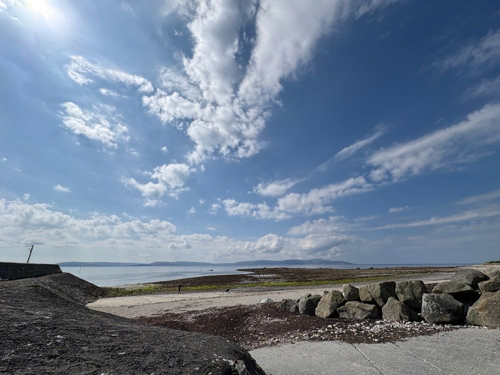 Dramatic cloudscape over a rocky beach shoreline with white puffy clouds stretching across bright blue sky.