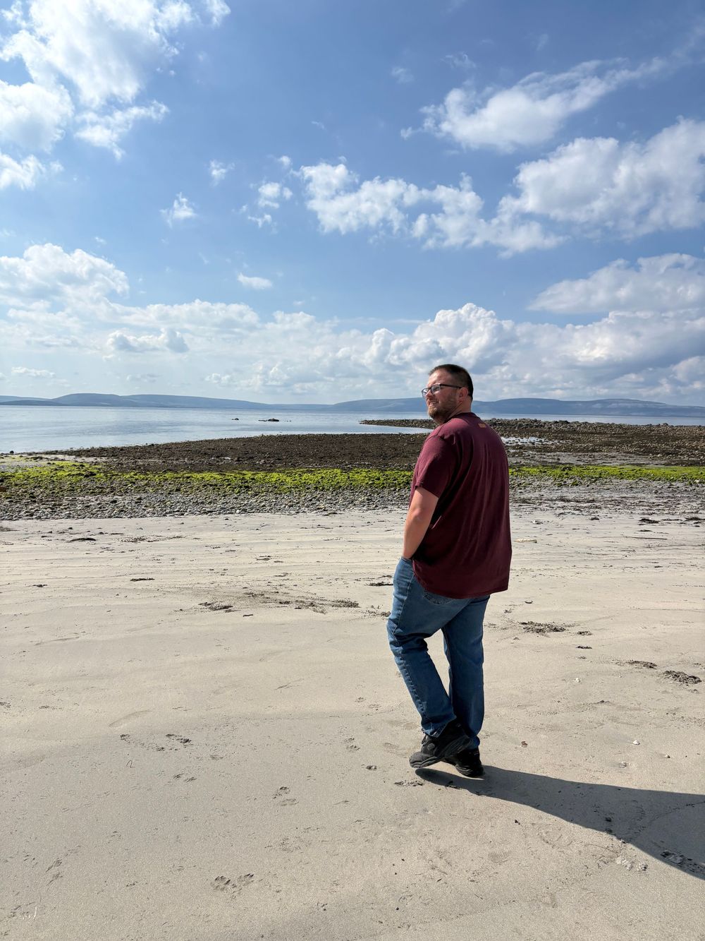Person in a red shirt and jeans standing on a scenic beach with water and cloudy blue sky in the background.