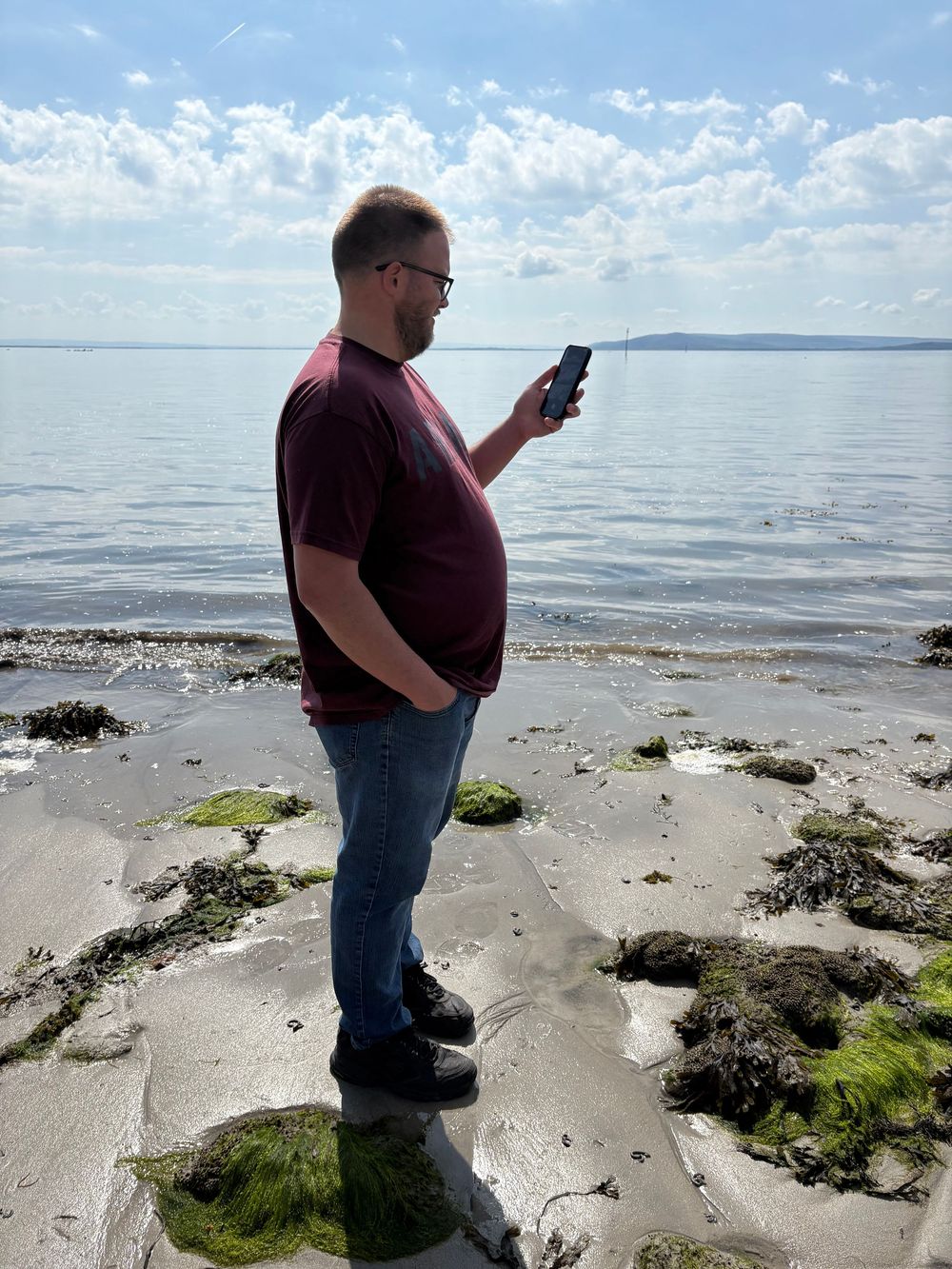 Person in casual attire checking phone while standing on algae-covered beach shore with calm water and cloudy sky.
