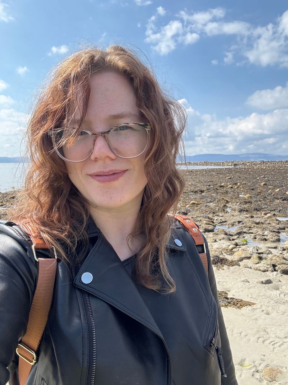 Selfie on a rocky beach with clear blue sky and water in the background, wearing glasses and a leather jacket.