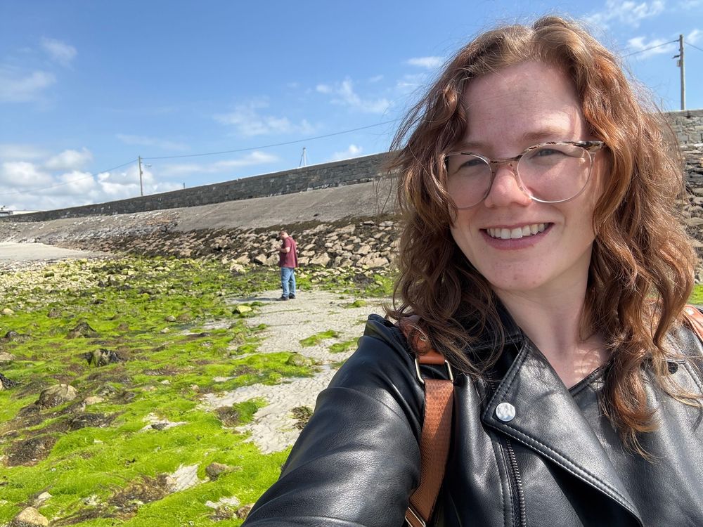 A person takes a selfie while hiking on a rocky trail with green moss on a bright sunny day with blue skies.