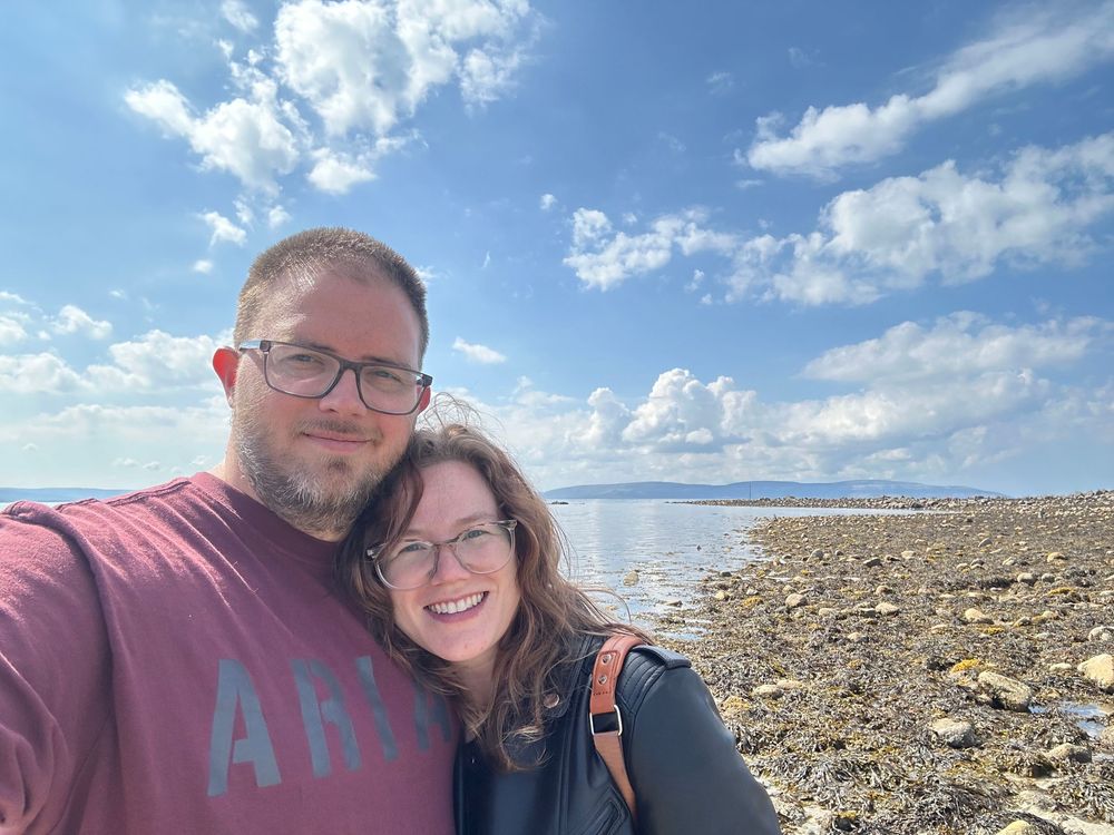 A couple takes a seaside selfie on a rocky beach shore under bright blue skies with puffy white clouds.