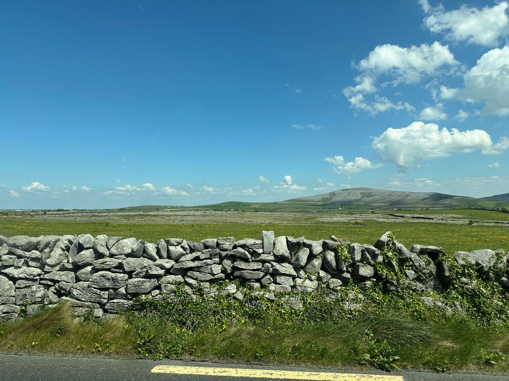 Stone wall along a rural road with green hills and mountains in the distance under a blue summer sky with white clouds.