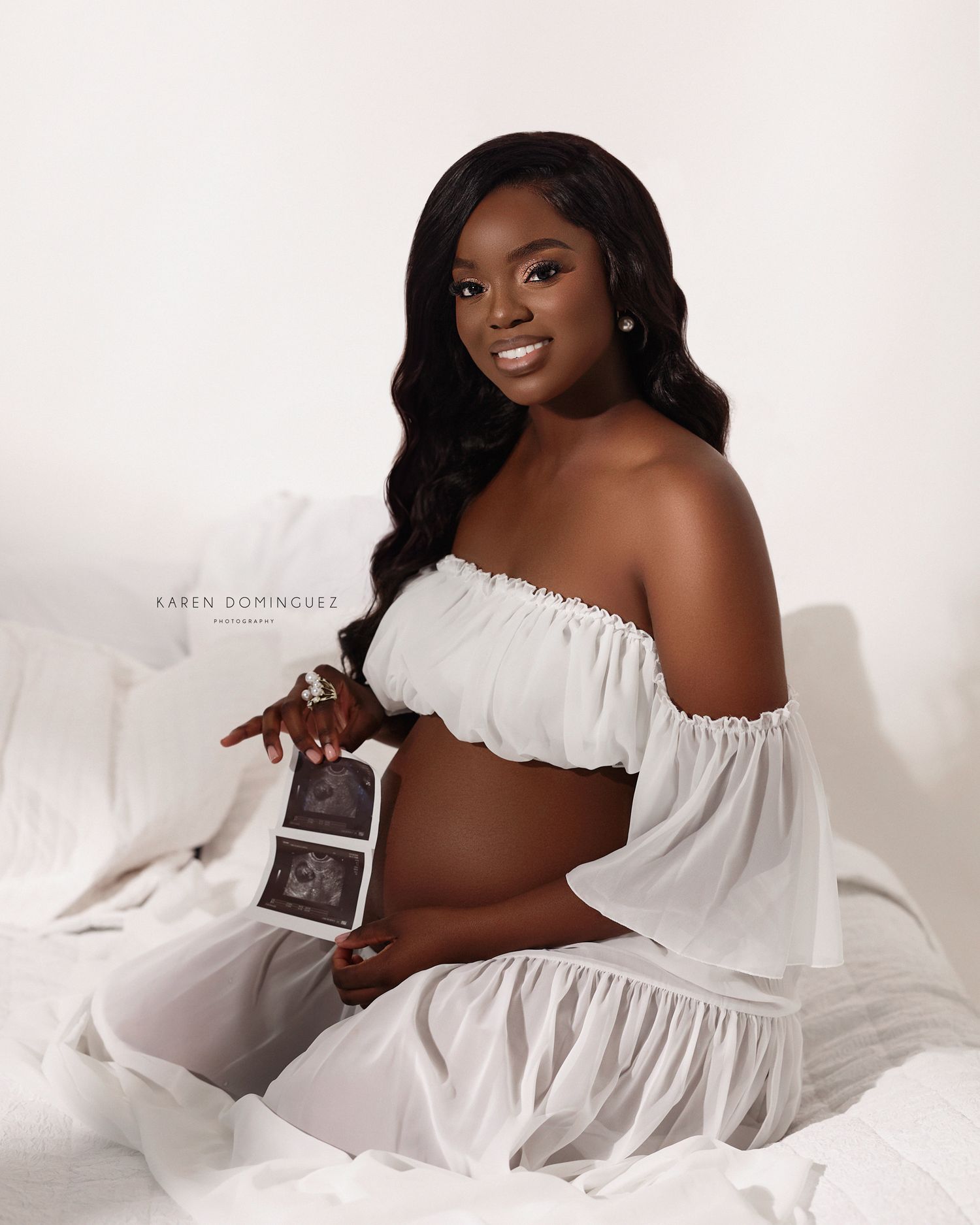 Beautiful maternity photo session in white off-shoulder dress against light background showing ultrasound images.