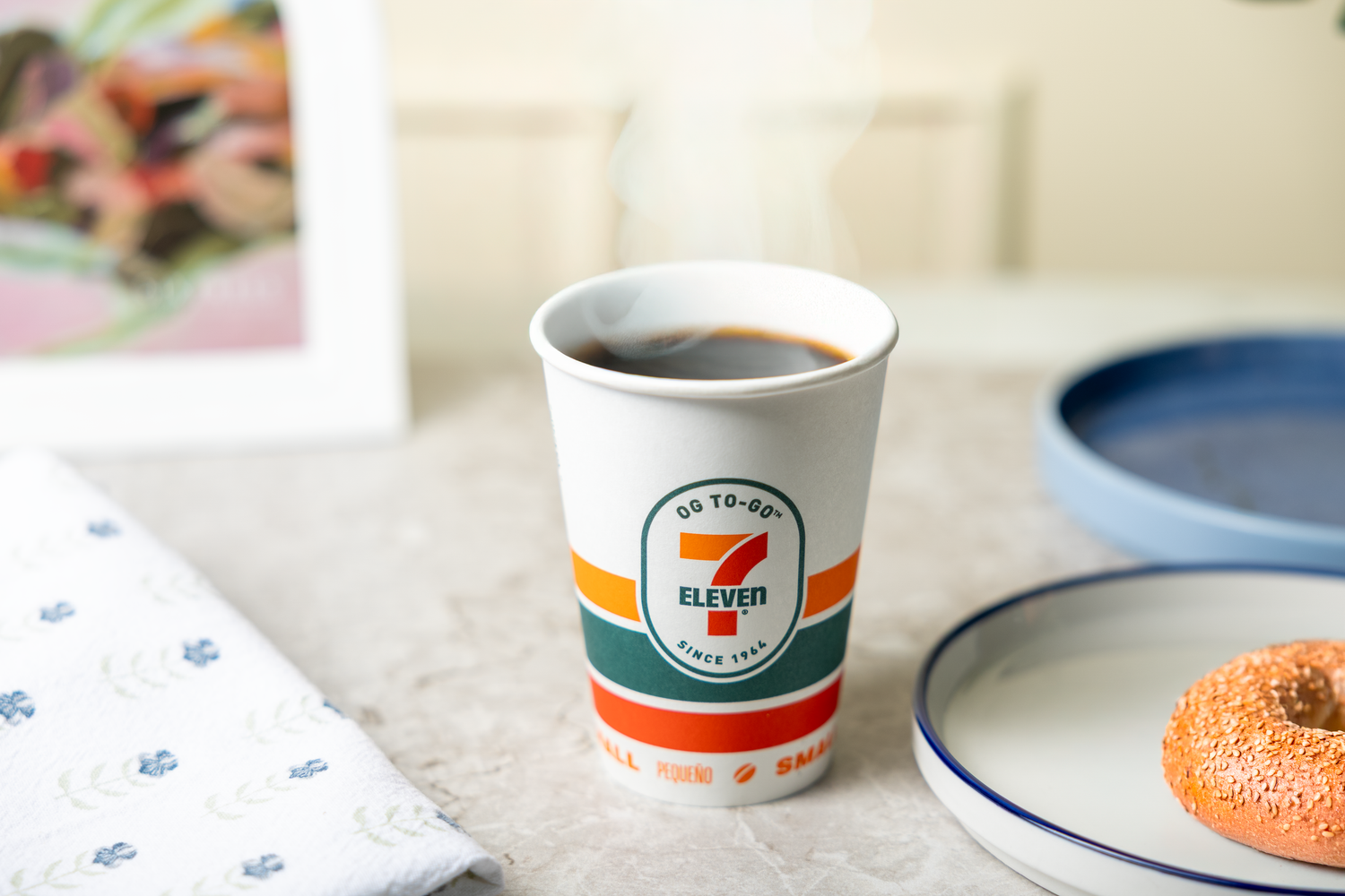 A 7-Eleven paper coffee cup sits on a white surface next to a blue-rimmed plate.
