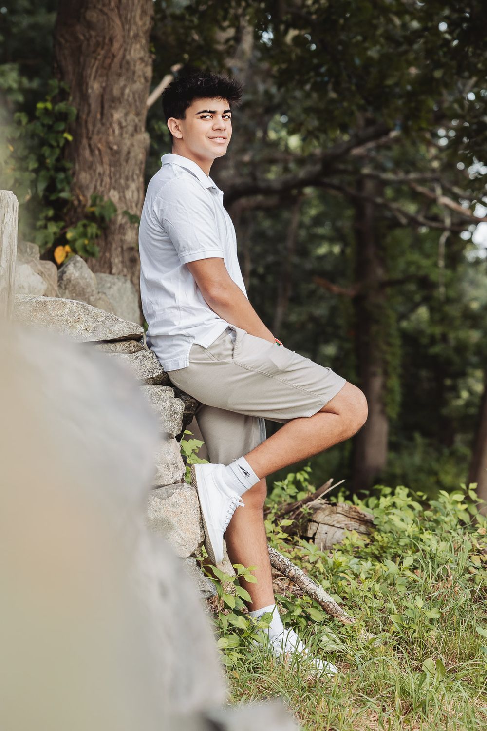 Young person in white polo shirt and khaki shorts sits casually on stone wall in woodsy outdoor setting.